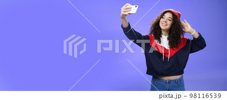 Cute girl making selfie to memorize trip standing in warm winter beanie and sweatshirt posing as pulling hand with smartphone and smiling at device screen to take pretty shot and post internet Cute girl making selfie to memorize trip standing in warm winter beanie and sweatshirt posing as pulling hand with smartphone and smiling at device screen to take pretty shot and post internet 98116539
