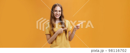 Girl shows cool place to visit. Portrait of charming carefree pretty young lady with cute natural wavy hair pointing left with index fingers and smiling broadly howing perfect place for advertisement 98116702