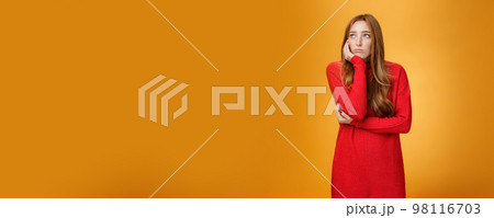 Sorrow and upset lonely woman leaning head on palm in upset pose looking gloomy at upper left corner as remembering unhappy story or being bored standing uneasy over orange wall 98116703