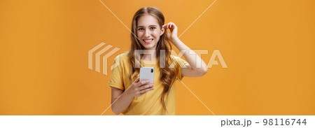 Waist-up shot of cute and friendly-looking nice young girl with wavy hairstyle putting hair strand behind ear holding smartphone smiling broadly with joyful expression at camera over orange background 98116744