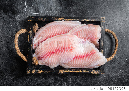 Fresh tilapia white fish fillet in a wooden tray. Black background. Top view Fresh tilapia white fish fillet in a wooden tray. Black background. Top view 98122303