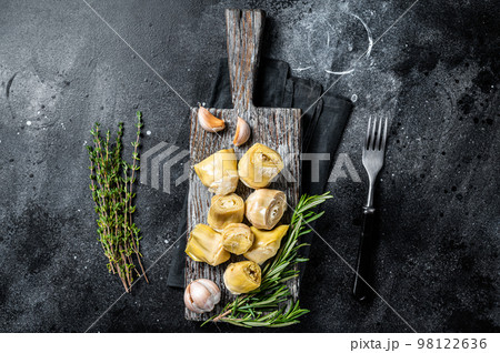 Artichokes hearts marinated with olive oil and herbs, pickled artichoke with garlic on wooden board. Black background. Top view 98122636