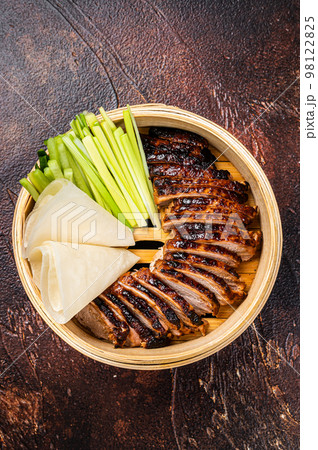Peking Duck in bamboo steamer served with cucumber, green onion, and wheaten chinese pancakes. Dark background. Top view 98122825