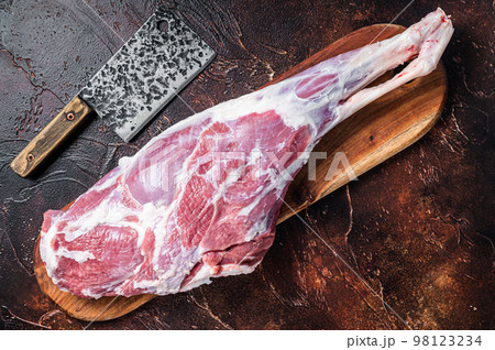Raw whole lamb leg thigh on butcher board. Dark background. Top view Raw whole lamb leg thigh on butcher board. Dark background. Top view 98123234