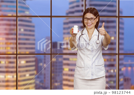 Young female doctor in glasses is showing medicine bottle and giving thumb up. Checkered windows with night cityscape view. Young female doctor in glasses is showing medicine bottle and giving thumb up. Checkered windows with night cityscape view. 98123744