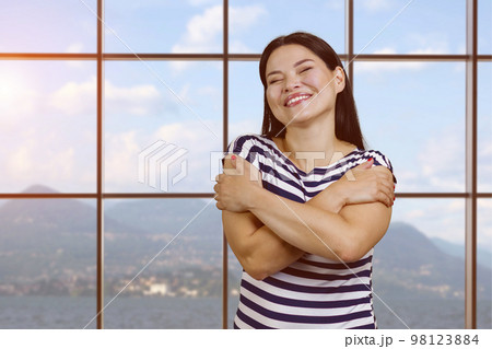 Portrait of happy young asian woman hugging herself. Checkered windows with view on mountain landscape on background. Portrait of happy young asian woman hugging herself. Checkered windows with view on mountain landscape on background. 98123884