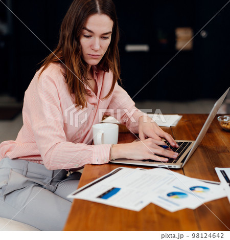 Young woman working with documents and laptop in the kitchen at home. 98124642