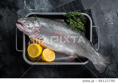 Raw whole trout fish in a kitchen tray with herbs. Black background. Top view 98125790