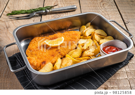 Pork meat weiner schnitzel with potato wedges and tomato sauce in steel tray. Wooden background. Top view 98125858