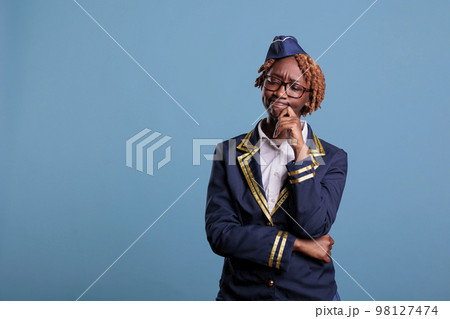 African american female flight attendant with doubtful expression wearing uniform against blue background in studio shot. Flight attendant thinking about the best in flight service strategy. 98127474