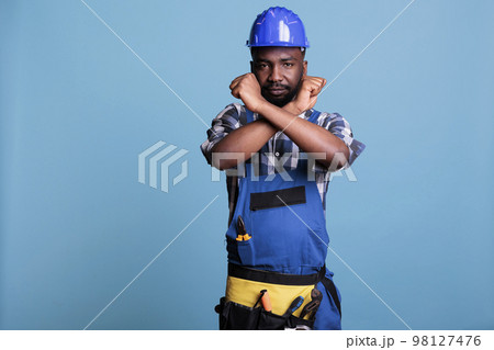 Disapproving african american builder making stop gesture with crossed arms while standing on blue background. Construction worker showing deny hands symbol while refusing to accept more work. 98127476