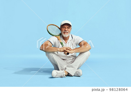 Delightful. Portrait of handsome senior man in stylish white outfit sitting, posing with tennis racket over blue background. Concept of leisure activity, hobby, lifestyle Delightful. Portrait of handsome senior man in stylish white outfit sitting, posing with tennis racket over blue background. Concept of leisure activity, hobby, lifestyle 98128064