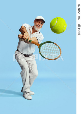 Returning ball. Portrait of handsome senior man in stylish white outfit playing tennis over blue background. Concept of leisure activity, hobby, lifestyle Returning ball. Portrait of handsome senior man in stylish white outfit playing tennis over blue background. Concept of leisure activity, hobby, lifestyle 98128076