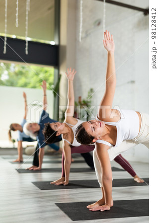 Calm young woman practicing Extended Side Angle Pose Utthita Parsvakonasana during group yoga class in studio Calm young woman practicing Extended Side Angle Pose Utthita Parsvakonasana during group yoga class in studio 98128412