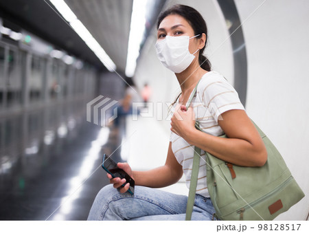 Woman in mask with smartphone sitting on bench in subway station Woman in mask with smartphone sitting on bench in subway station 98128517