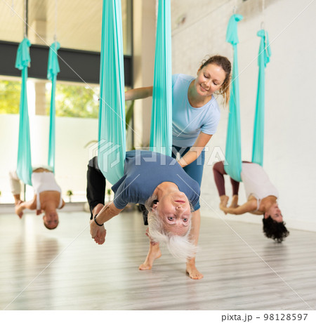 Supportive female trainer helping to mature female during antigravity aerial yoga exercise in studio 98128597