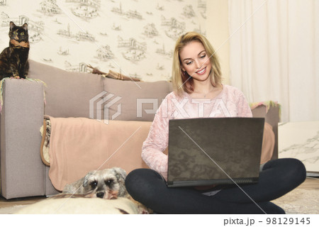 Lady smiles during a video call on a laptop, little cat sitting on the couch and cute dog is sleeping on a pillow nearby. 98129145