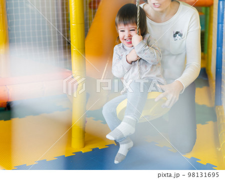 Toddler plays on rope swing with his mother or babysitter. Physical development for little children. Interior of kindergarten or nursery. Toddler plays on rope swing with his mother or babysitter. Physical development for little children. Interior of kindergarten or nursery. 98131695