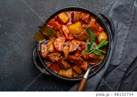 Beef meat stew with potatoes, carrot and delicious gravy in black casserole pot with bay leaves and fresh green herbs with spoon on black dark rustic concrete background from above . Beef meat stew with potatoes, carrot and delicious gravy in black casserole pot with bay leaves and fresh green herbs with spoon on black dark rustic concrete background from above . 98132754