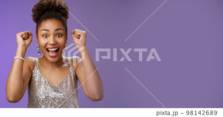 Happy astonished african american woman in silver fashionable dress winning first prize gladly celebrating raising clenched fists dancing triumphing happily having fun, standing blue background 98142689