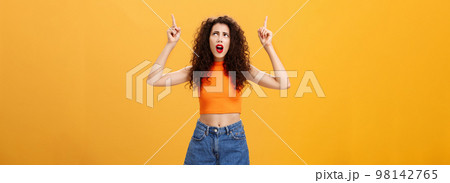 Silly and moody european party girl with curly hairstyle in red lipstic and stylish orange cropped top complaining on bad weather raising hands pointing and looking up with displeased unhappy face Silly and moody european party girl with curly hairstyle in red lipstic and stylish orange cropped top complaining on bad weather raising hands pointing and looking up with displeased unhappy face 98142765