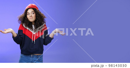 Not getting clue. Cute silly woman with curly hair shrugging with pursed lips and unbothered expression holding hands sideways looking right being unaware and confused, uncertain what answer 98143030