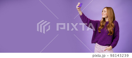 Attractive confident and stylish happy 20s woman with ginger hair standing carefree with joyful smile over purple background extending hand with smartphone as taking selfie happily Attractive confident and stylish happy 20s woman with ginger hair standing carefree with joyful smile over purple background extending hand with smartphone as taking selfie happily 98143239