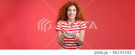 Excited happy cute redhead curly-haired girlfriend eager start learn how play boyfriend game look amused cheerful camera smiling joyful hold horizontal smartphone load app red background 98143392