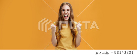 Portrait of happy cheerful lucky young european girl with long natural wavy hair raising clenched fists in cheer and joy yelling yes from triumph feeling glorious moment of win over orange wall 98143801