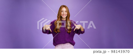 Girl giving recommendation to visit place, pointing down and smiling friendly with delighted and pleasant grin. Portrait of attractive redhead female model in purple warm sweater promoting copy space 98143970