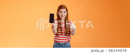 Impressed excited happy redhead woman smiling broadly, hold smartphone, pointing cellphone screen, thrilled amazed introduce fascinating application, stand orange background rejoice Impressed excited happy redhead woman smiling broadly, hold smartphone, pointing cellphone screen, thrilled amazed introduce fascinating application, stand orange background rejoice 98144076