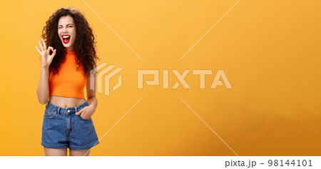 Excellent party like it. Joyful enthusiastic young curly-haired female with red lipstick in stylish cropped top winking excited smiling and showing okay or perfect sign posing over orange background Excellent party like it. Joyful enthusiastic young curly-haired female with red lipstick in stylish cropped top winking excited smiling and showing okay or perfect sign posing over orange background 98144101