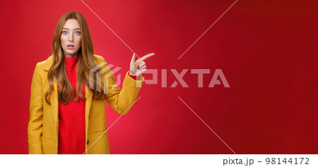 Impressed and questioned amazed redhead woman remain speechless from amazement pointing right open mouth in stupor, asking question about confusing shocking promotion over red wall 98144172