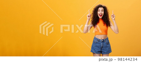 Woman surprising boyfriend with cool sign hanging upstairs pointing up, smiling singing happy b-day standing carefree and joyful over orange background in stylish cropped top and summer denim shorts Woman surprising boyfriend with cool sign hanging upstairs pointing up, smiling singing happy b-day standing carefree and joyful over orange background in stylish cropped top and summer denim shorts 98144234
