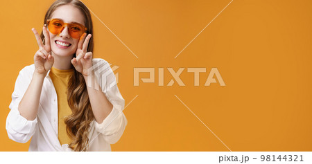 Stylish enthusiastic and charismatic young party girl in trendy sunglasses and white blouse over t-shirt showing peace gestures near face and smiling cute at camera against orange background Stylish enthusiastic and charismatic young party girl in trendy sunglasses and white blouse over t-shirt showing peace gestures near face and smiling cute at camera against orange background 98144321