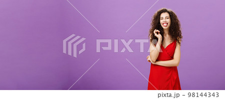 Studio shot of playful and joyful artistic caucasian female student in red evening dress sticking out tongue happily having fun playing with curl standing over purple background careless and carefree 98144343