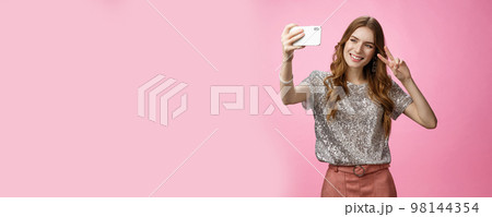 Sociable attractive glamour young caucasian woman taking selfie show victory peace sign smiling broadly raise smartphone look grinning mobile phone display, video calling friends, pink background 98144354