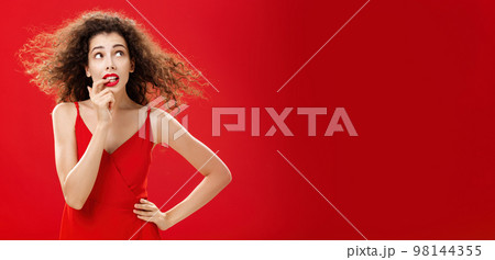 Waist-up shot of concerned and worried insecure cute female with curly hairstyle in red evening dress biting finger looking troubled or questioned at upper right corner with hand on waist 98144355