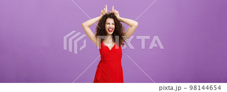 Devil lives inside lady. Daring stylish adult woman with curly hairstyle in red evening dress winking making confident and amused expression showing horns with index fingers on head, being stubborn 98144654