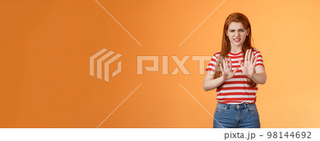 Gosh it stinks. Disgusted redhead picky woman blocking sign raise hands up defensive, grimacing, cringe from aversion awful smell, show refusal rejecting disgusting offer, stand orange background 98144692