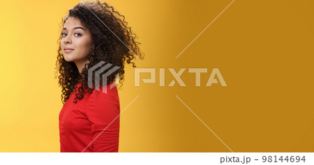 Profile shot of stylish and cool attractive curly-haired european 25s woman in red dress turning at camera and smiling broadly with delighted carefree expression, having fun over yellow background Profile shot of stylish and cool attractive curly-haired european 25s woman in red dress turning at camera and smiling broadly with delighted carefree expression, having fun over yellow background 98144694