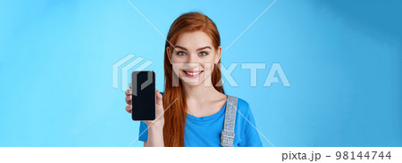 Close-up attractive joyful redhead young 20s female shop assitant suggest customer buy smartphone, show phone display, promote application, internet app, smiling camera, stand blue background 98144744