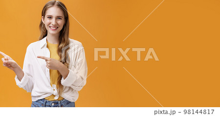 Waist-up shot of friendly pleasant and stylish young teenage female in blouse over t-shirt with cute wavy fair hair pointing left and smiling broadly at camera posing happy against orange background 98144817
