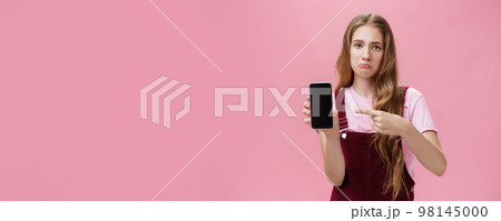 Sad gloomy young female with cute wavy natural hairstyle showing smartphone screen pointing at gadget with index finger making upset face frowning displeased feeling regret after buying broken phone Sad gloomy young female with cute wavy natural hairstyle showing smartphone screen pointing at gadget with index finger making upset face frowning displeased feeling regret after buying broken phone 98145000