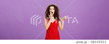 Portrait of surprised and amazed charming elegant lady with evening make-up in red dress covering opened mouth from shock and amazement pointing left curious and questioned over purple background Portrait of surprised and amazed charming elegant lady with evening make-up in red dress covering opened mouth from shock and amazement pointing left curious and questioned over purple background 98145021