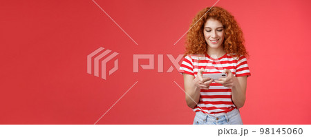 Redhead girl fool around waiting queue dentist playing awesome smartphone game hold phone horizontal tap cellphone screen look telephone display smiling delighted entertained standing red background Redhead girl fool around waiting queue dentist playing awesome smartphone game hold phone horizontal tap cellphone screen look telephone display smiling delighted entertained standing red background 98145060