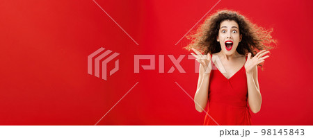 Portrait of surprised and astonished speechless curly-haired woman in red luxurious dress dropping jaw from amazement popping eyes at camera and gesturing emotive and shocked over background 98145843