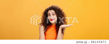 Well sorry. Unaware and clueless silly charming woman with curly hairstyle shrugging and smirking with uncertain look raising palm in who knows gesture, posing uninvolved over orange background 98145871