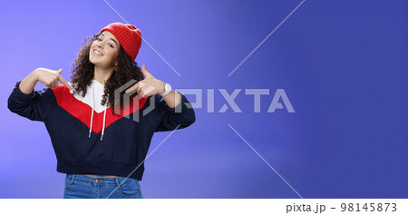 Proud and self-satisfied happy young energized stylish female model in warm beanie brag about own achievements as pointing at herself raising head and smiling produly posing delighted over blue wall 98145873