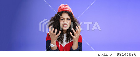 Pissed and annoyed stylish girl clenching hands in anger, grimacing squeezing teeth from annoyance and irritation, standing hateful and outraged feeling fury and hate posing over blue background 98145936